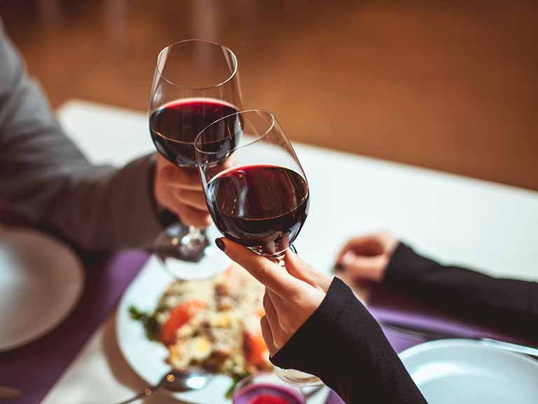 Two people clinking glasses of red wine over a table with food.