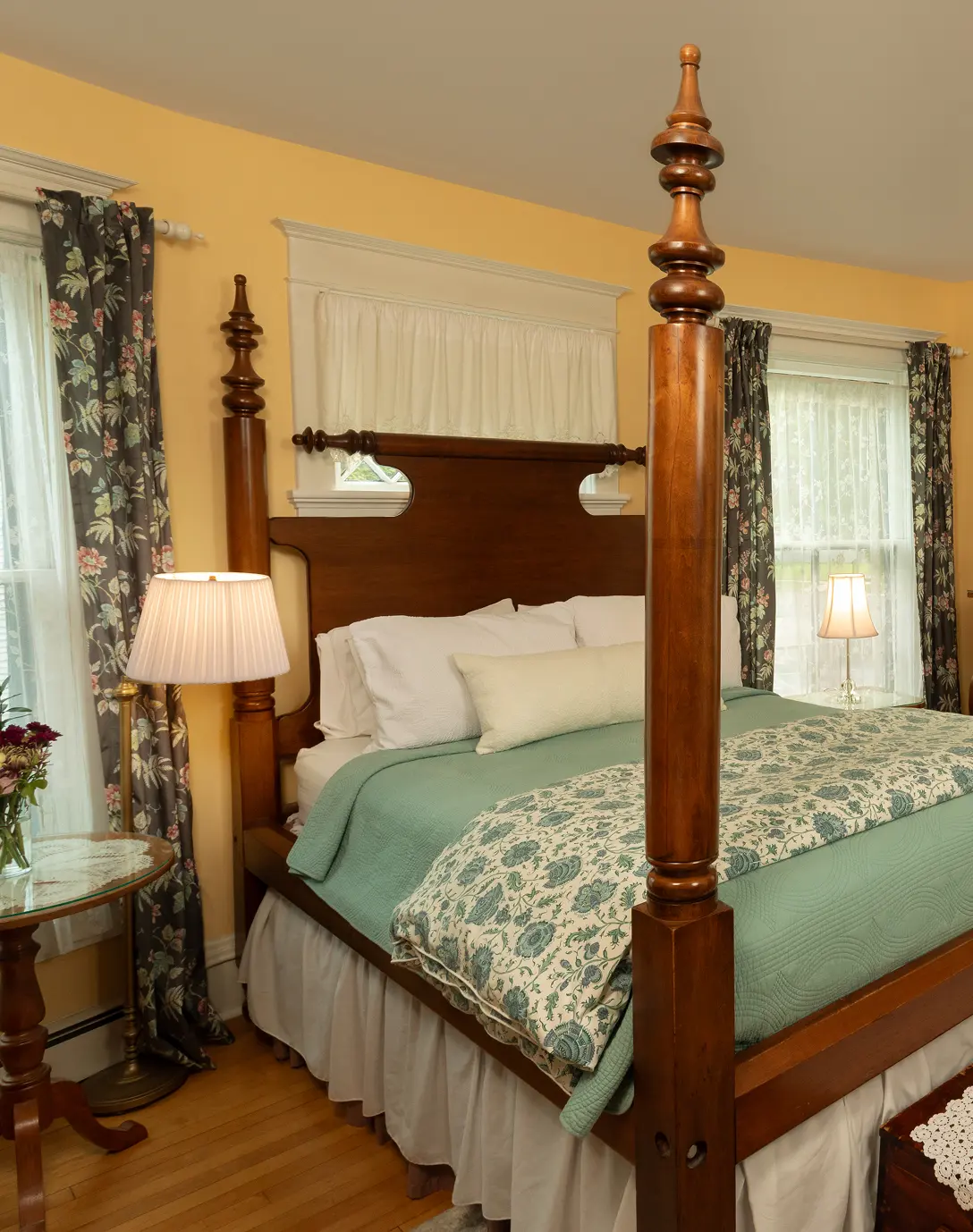 A bedroom with a wooden four-poster bed, mint green bedding, floral curtains, and yellow walls.