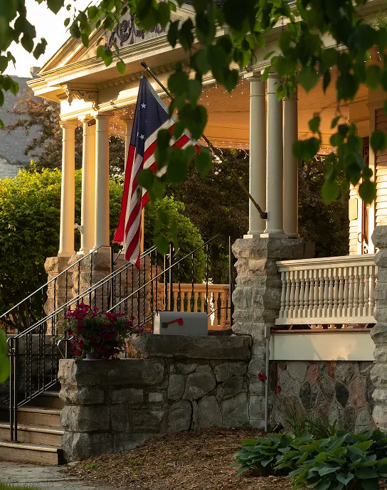 A historic home’s porch with white columns, a fieldstone base, and an American flag hanging from a pole.
