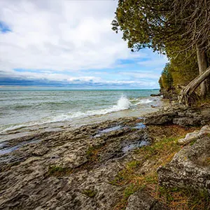 Waves crash against a rocky, tree-lined shoreline under a cloudy, blue sky.