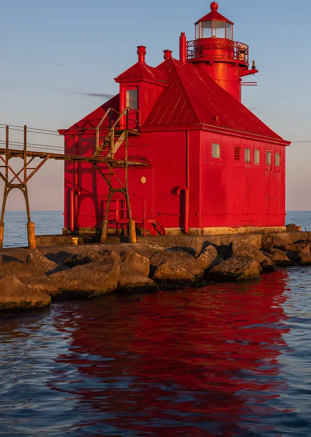 The vibrant red Algoma Pierhead Lighthouse sits at the end of a rocky breakwater, reflected in the calm water at sunset.