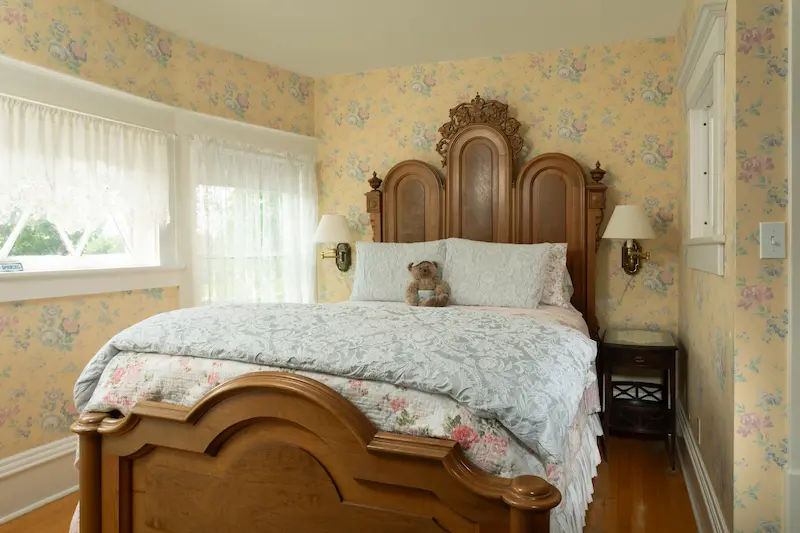 A bedroom with yellow floral wallpaper, a wooden bed frame, a teddy bear, and white curtains.