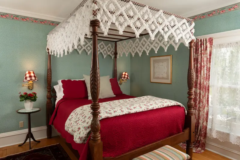 A four-poster bed with a white lace canopy in a bedroom with sage green walls and red accents.