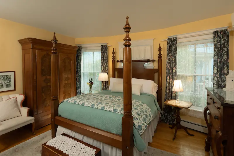 A furnished bedroom with a four-poster bed, wood furniture, yellow walls, and windows with patterned curtains.