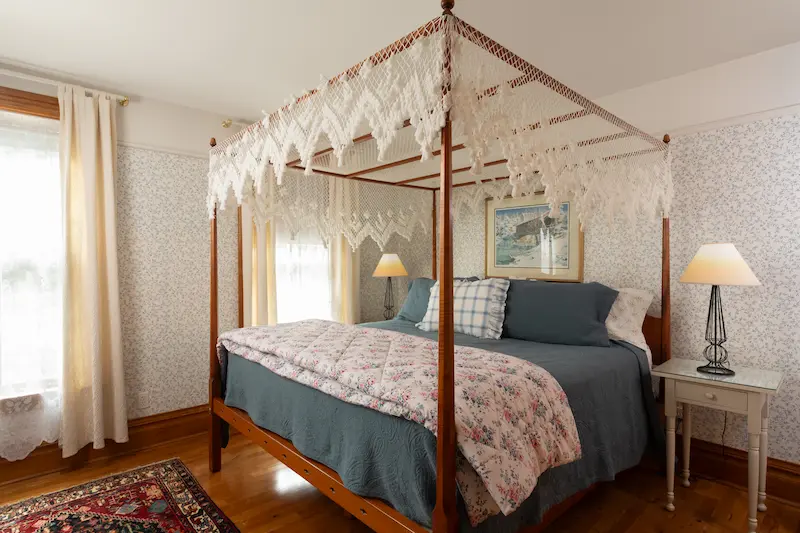 A wooden canopy bed with a macramé valance, dressed in blue and floral bedding in a room with patterned wallpaper.
